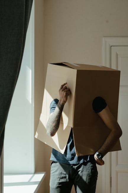 A person with tattoos wearing a casual grey t-shirt and black wristwatch is seen inside a room, carrying a large cardboard box over their head during a home relocation process. The individual's left a