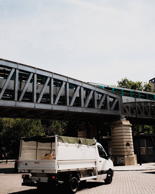 A small white cargo van parked on a paved area underneath a railway bridge during daytime, with a green tarpaulin partially covering an open cargo area containing cardboard boxes. The bridge is constr