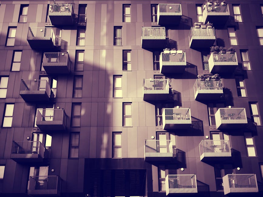 Photograph of a tall multi-storey residential building seen from ground level, looking upward towards the blue sky with scattered white clouds. The building features numerous windows with white frames