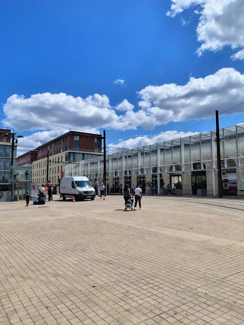 A spacious outdoor area with a paved surface featuring rectangular tiles, adjacent to a modern building with large glass windows and a metallic framework. Several people are present, some standing and