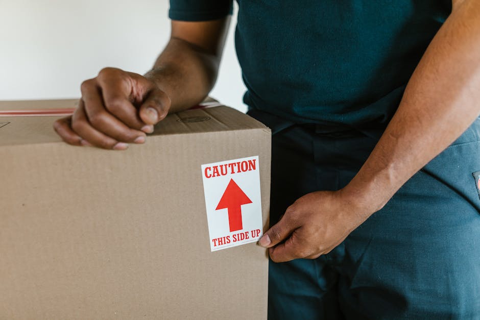 A man with a beard wearing a white polo shirt and a black cap is standing outside during daylight, holding a cardboard box near the back of a moving van. The van's open rear doors reveal a loading are
