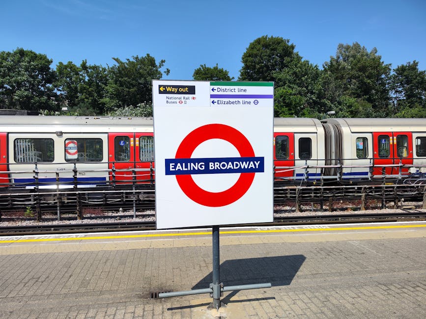 A railway station platform in London with a prominent Ealing Broadway sign mounted on a metal stand in the foreground. Behind the sign, there is a modern train with a silver body, red and blue accents